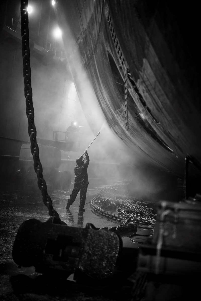 Svartvitt fotografi, där en person står i dimman och sprutar vatten på skrovet på ett fartyg i en torrdocka.