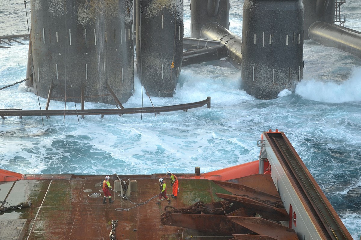 Three men working at the edge of an anchor handling vessel. Behind them a large platform in aggressive  water