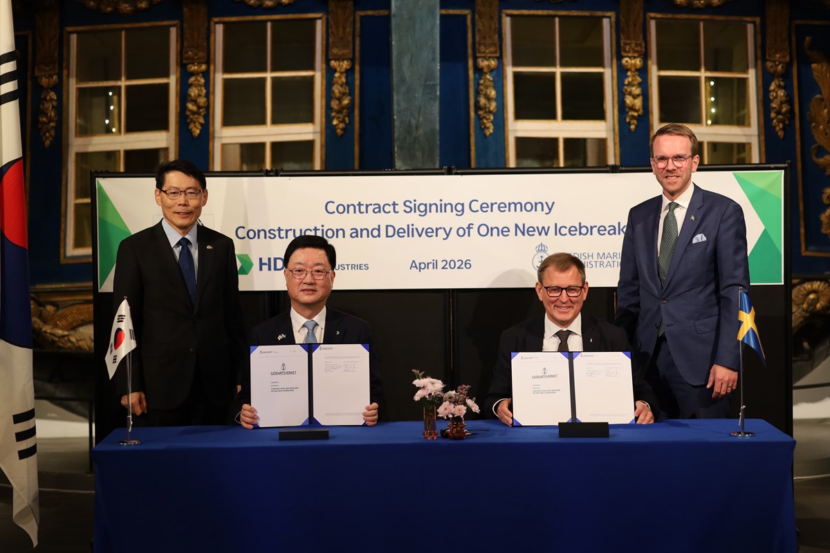 Fyra män, två sitter och visar upp ett kontrakt. De andra två står vid sidan av. Alla tittar in i kameran och ler.