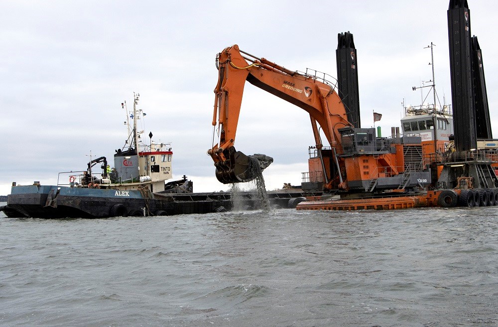 Arbetet inleds på den nordvästra sidan av muddringsområdet Kälsholmen samt vid Galtgrynnan. Foto: Agne Hörnestig. Muddringsverket i Hargs Hamn är igång. En bogserbåt syns intill, och grävskopan plockar upp muddermassa ur vattnet.