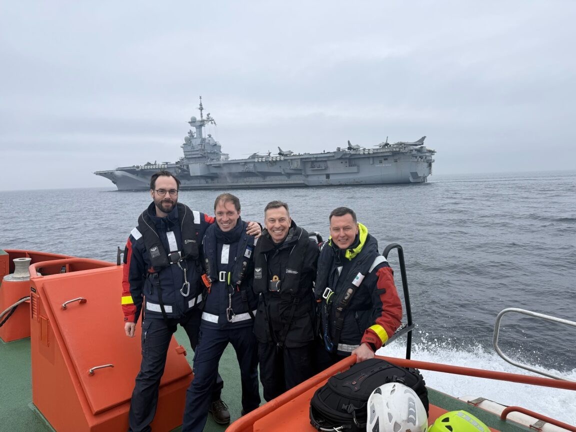 Hangarfartyg i bakgrunden. Lotsar står uppradade på en lotsbåt framför.