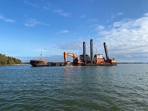 Vid mediebesöket pågick muddring vid Kälsholmen. Foto: Max Bjurström Muddringsverket i Kälsholmen, Hargs Hamn. Det är blå himmel och en grävskopa syns gräva under ytan.