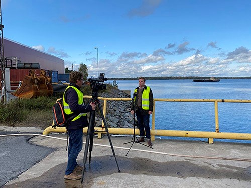 Projektledare Max Bjurström intervjuas av SVT Uppland. Foto: Hargs hamn Max Bjurström blir intervjuad framför vattnet. En kamera står uppställd framför honom, han tittar in i den. En reporter står bakom kameran. Det är soligt.