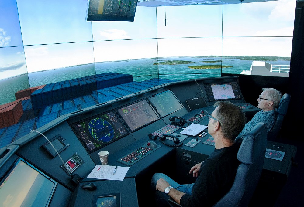 Inifrån simulatorn där stor skärmar av ett animerat hamninlopp syns, samt ett stort kontrollbord likt bryggan i ett fartyg.