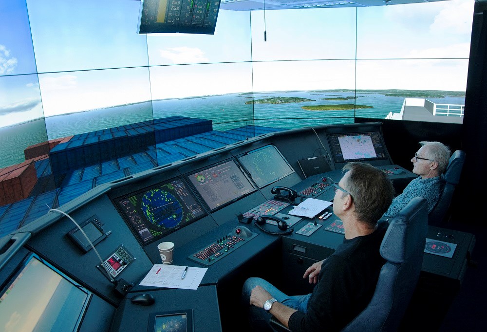 Containerfartyget som provkörs är 430 meter långt, 65 meter brett och fullastat. Den totala ytan motsvarar ett vindfång på drygt 20 000 kvadratmeter. Foto: Agne Hörnestig. Inifrån simulatorn där stor skärmar av ett animerat hamninlopp syns, samt ett stort kontrollbord likt bryggan i ett fartyg.