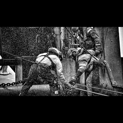 Black and white photo of fishermen in stormy weather handling anchor chain while attached to safety ropes