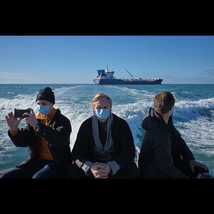 ”Signing off year” 2020 av Jörgen Språng. Tre sjömän med munskydd sitter i en mindre båt på väg ifrån ett större fartyg.