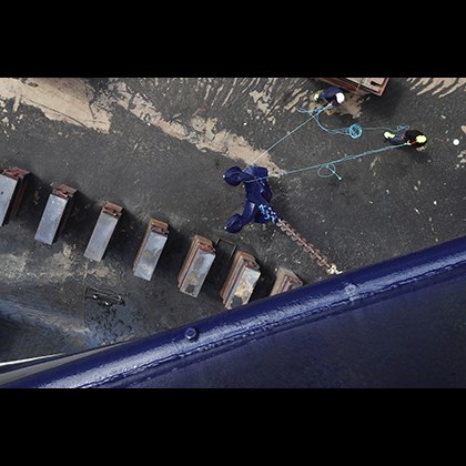 View from the gunwale. Two man handling a large anchor on the shipyard.