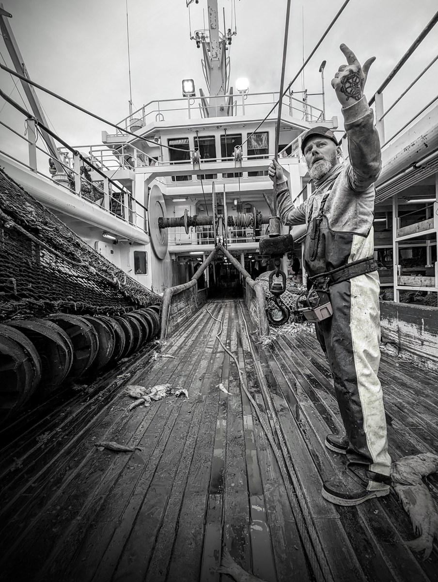 Black and whit photo of a fisher man with tattooed hand, waving to someone to come