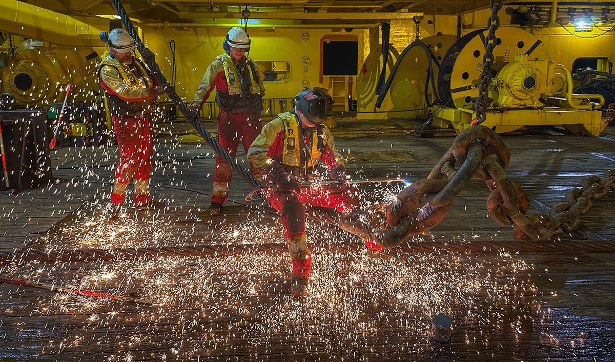 Three men are working with an anchor chain. Sparks are flying across the ship's deck