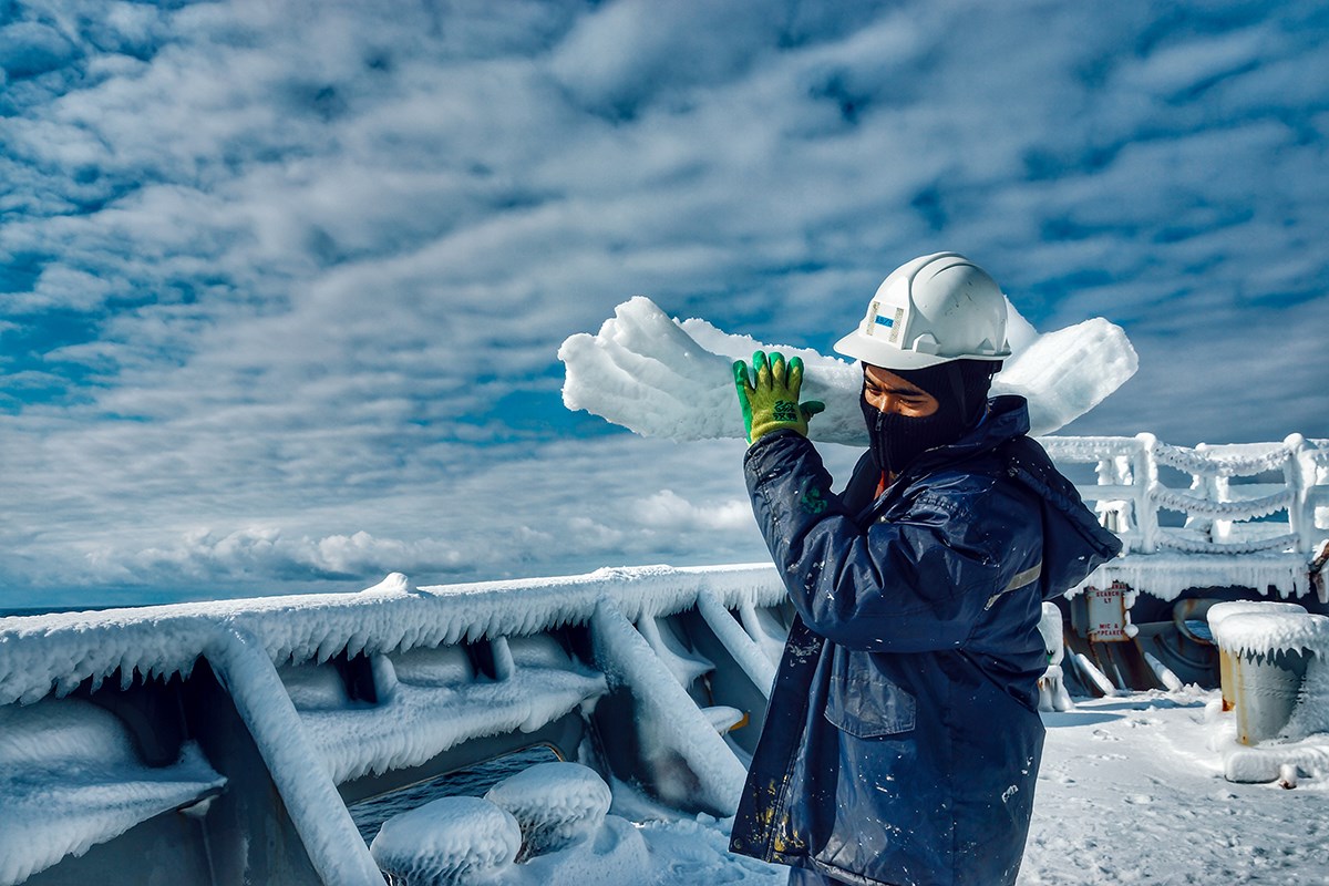 Sailor in a white helmet carrying a large chunk of ice aboard an iced ship
