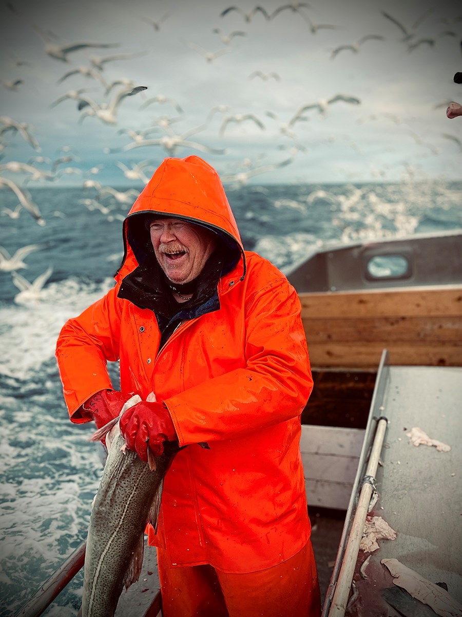 A fisherman in an orange raincoat holding a fish, with seagulls in the background
