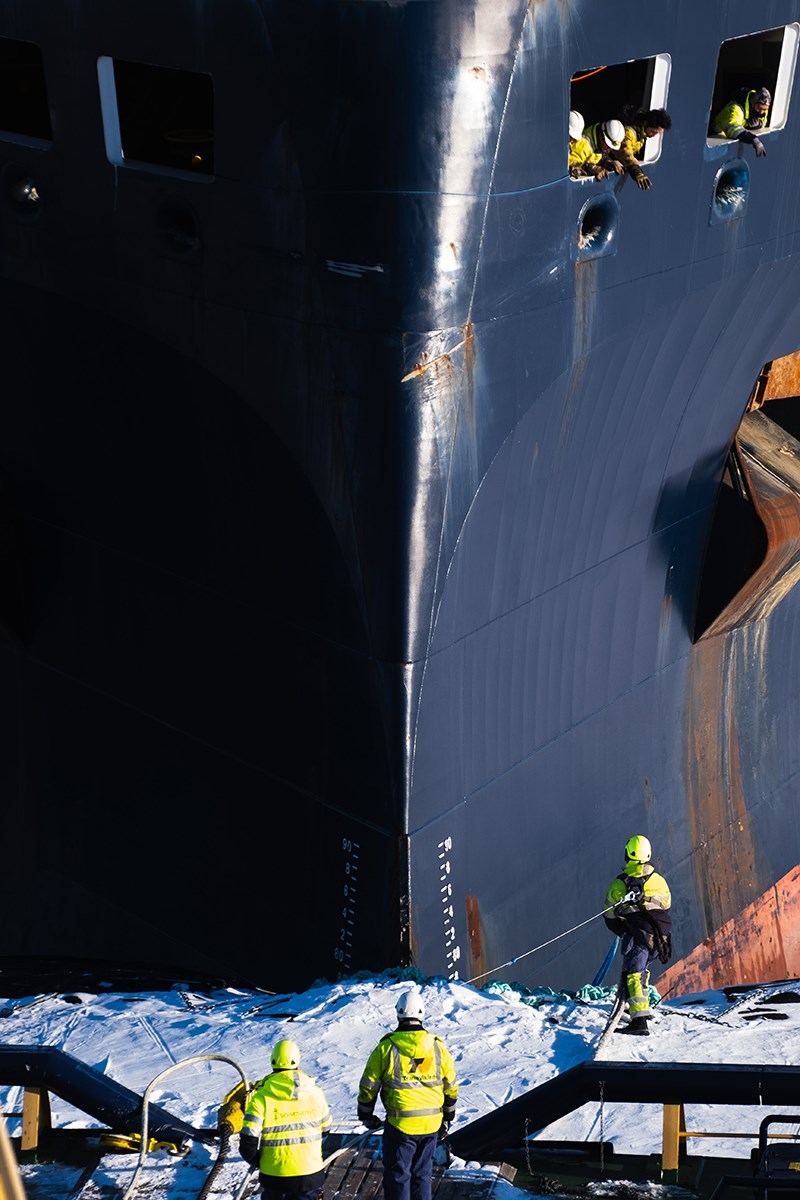 A ship's bow fills the image, some sailors are looking out of the anchor hatch and seem to be communicating with those standing at the bottom of the image
