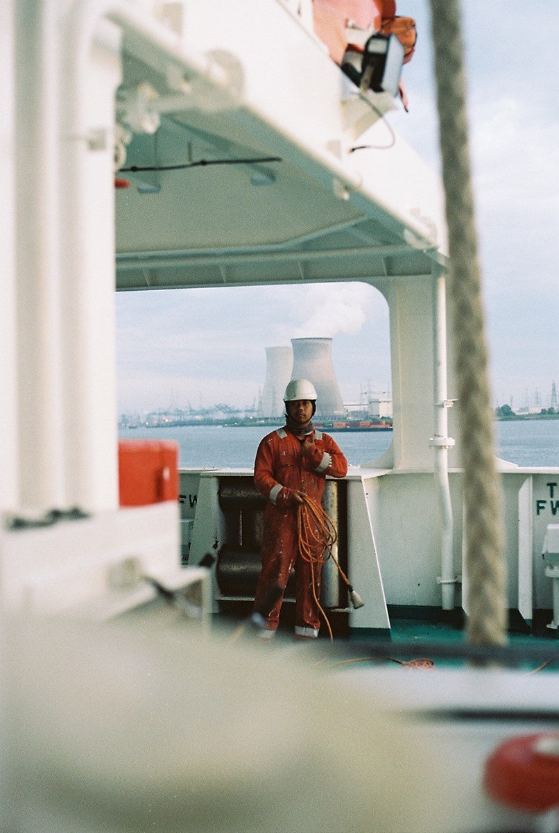 A sailor stands aboard his ship, dressed in overalls and holding an electric cable, with nuclear power plants visible in the background