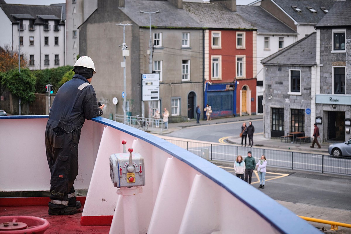 A ship's bow appears to almost hang over the harbor street in a village, a sailor looks into an Irish village, and villagers look back