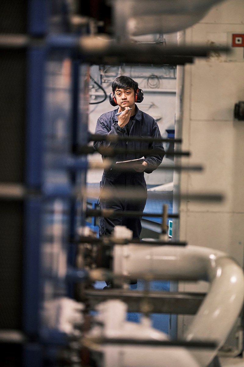 A sailor in overalls, ready to jot something down in the engine room, scratching his chin
