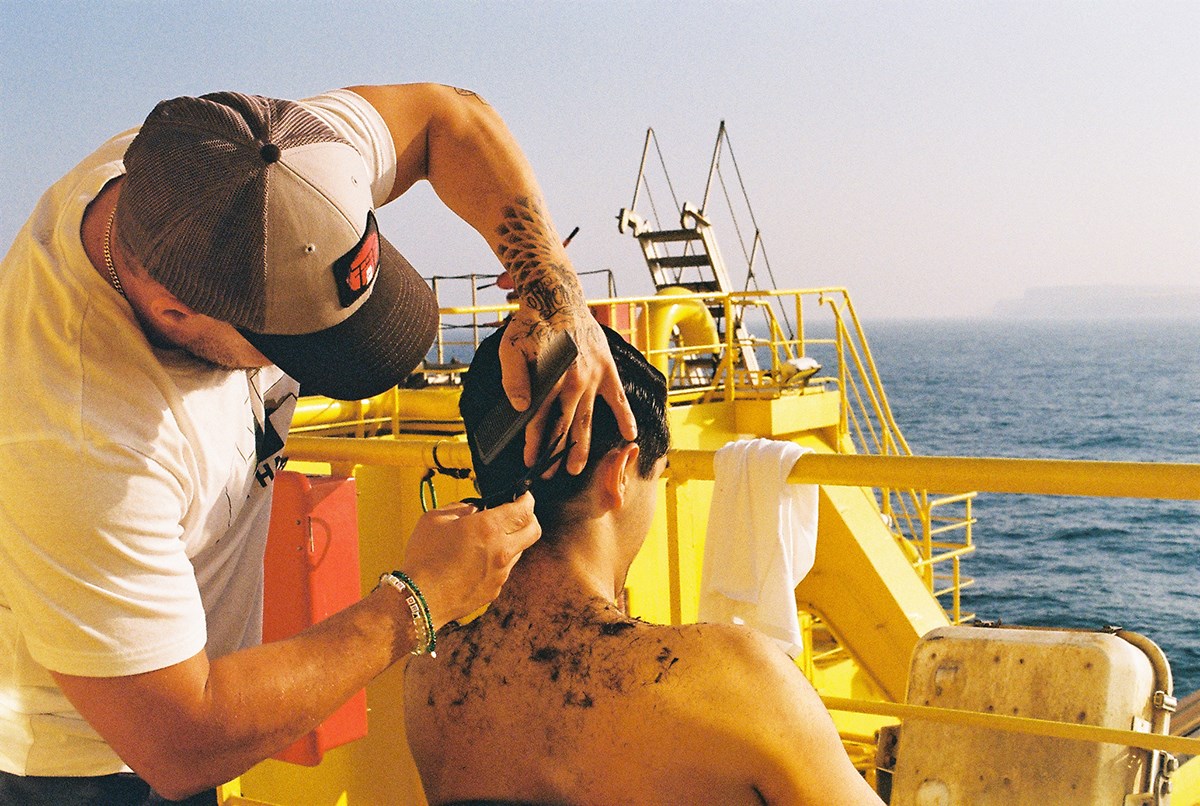 A man is cutting another man's hair on board, outdoors in the sunshine.