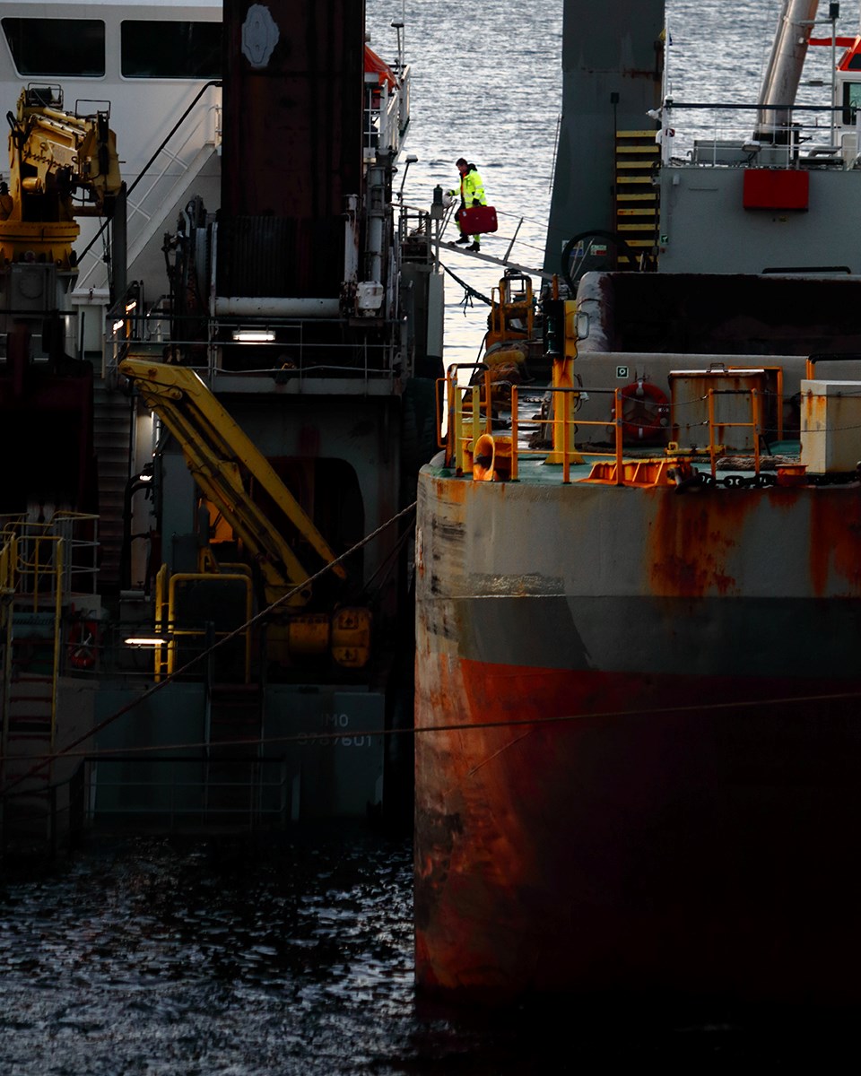 A man is walking on a gangway with something resembling a bag in his hand, between two ships