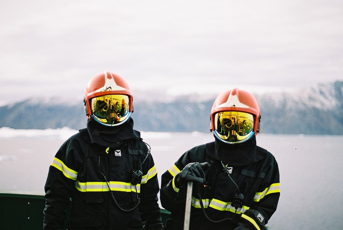 Two people dressed in firefighting gear and helmets with visors, looking into the camera. In the background, a mountainous landscape