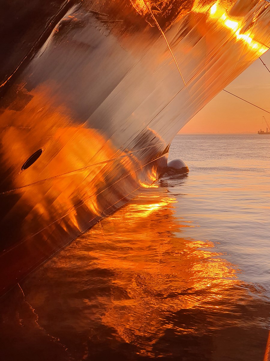A ship’s side and its reflection in the water, glowing in the morning sunlight