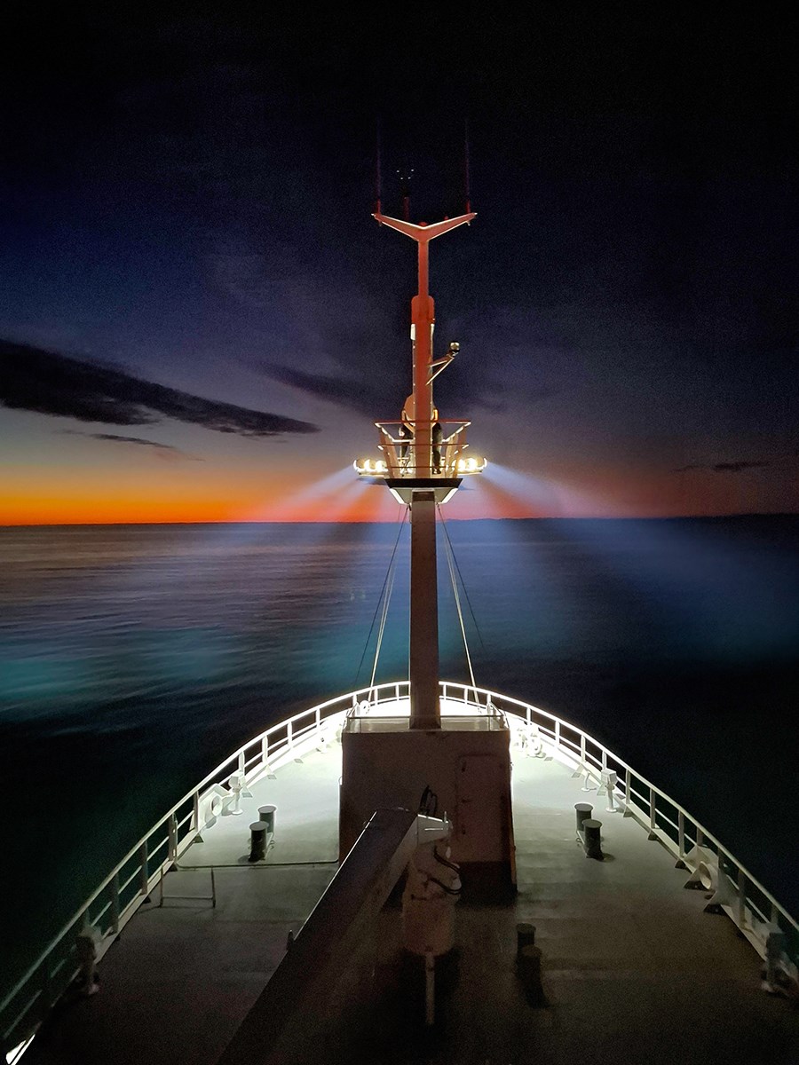 An illuminated ship’s bow moves toward the glowing horizon. The mast headlights make it glow as well. The colors of the water shift from turquoise to indigo