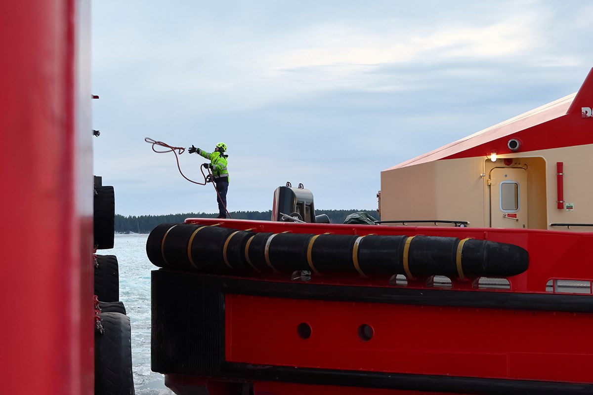 A man on a red ship throws a mooring line to another person, of whom only a hand is visible