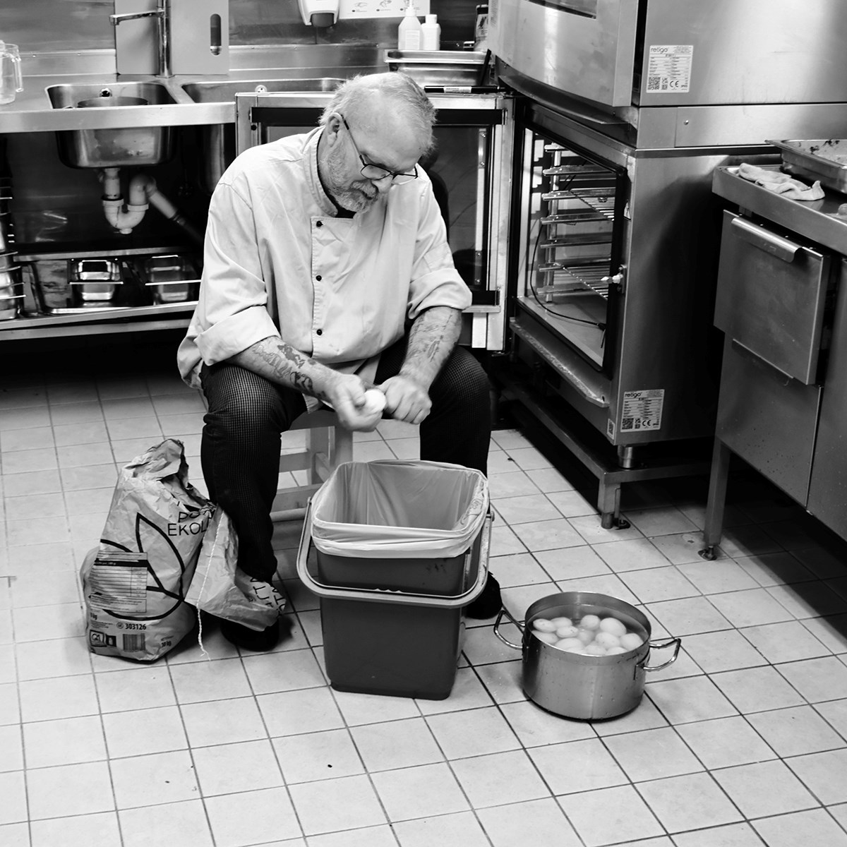 A black‑and‑white photo of a man in cook’s clothing sitting in the galley, peeling potatoes over a waste bin