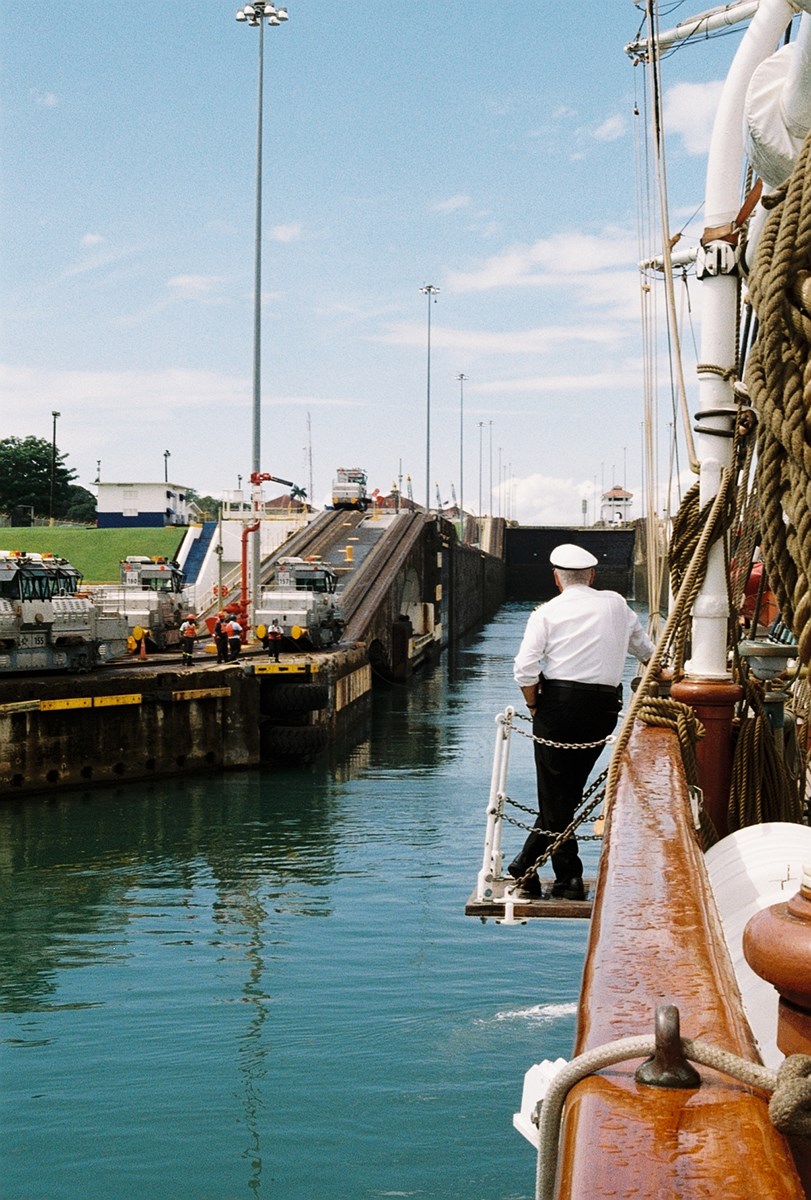 A sailing ship entering a lock. On the quay to the left, workers are visible. A man wearing a captain’s cap stands relaxed on the ship’s gangway, looking toward the lock gate.