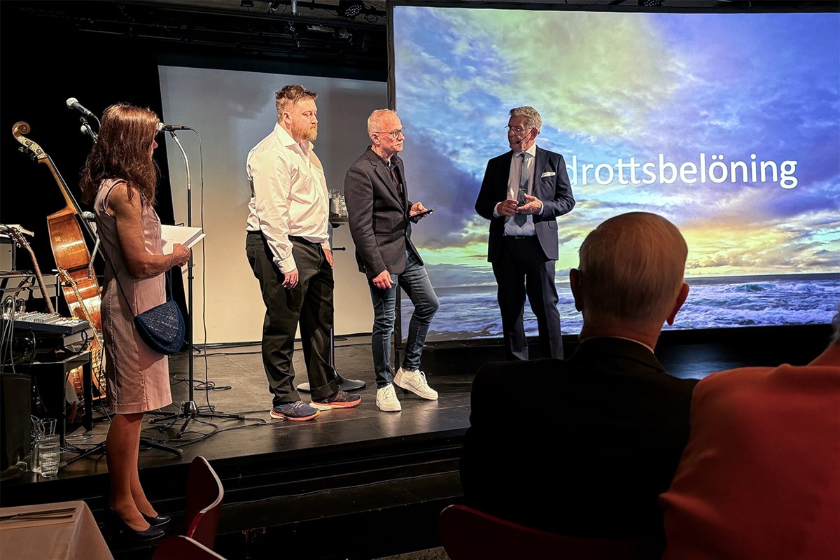 Utdelning av Idrottsbelöningen. Fr. vänster stiftelsens Birgitta Lönnquist Gemvik, representant från M/V Figaro, Glenn Hjelle och Karl Karell. Fyra personer står på en scen, i bakgrunden en projektorduk där det står Idrottsbelöning