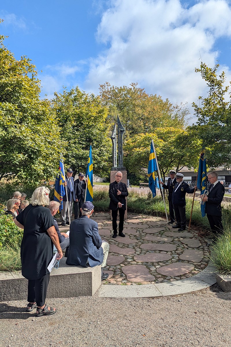 Åhörarna lyssnar till Mikael Billemars ord under minnesstunden i Norrköping. En grupp lyssnar till en präst som står och håller ett korum, vid ett minnesmärke. Några av åhörarna håller i flaggor.